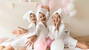 Three women in white bathrobes with towels on their heads smiling and holding pink and white balloons during a spa day for wedding party celebration.