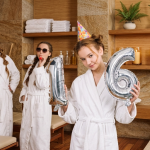Teenage girls celebrating a 16th birthday at a spa wearing white robes and party hats by a pool.